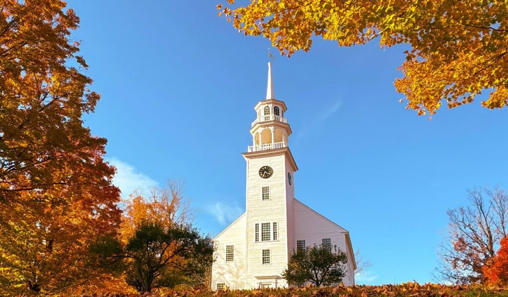 White church with a tall steeple and clock tower framed by vibrant autumn trees under a clear blue sky in Groton, Vermont