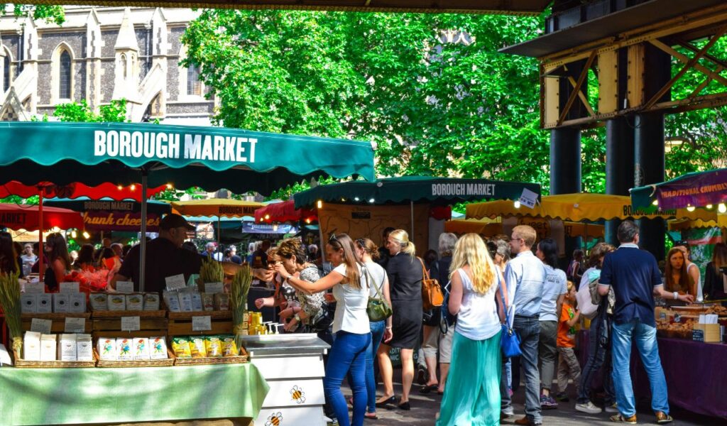 People browsing colorful stalls at an outdoor market, with green awnings reading "Borough Market" under leafy trees.
