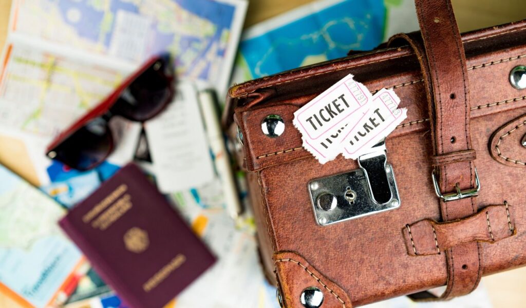 High Angle View of Tickets on top of Brown Leather Suitcase in Foreground with Passport, Sunglasses and Maps Scattered on Table in Background