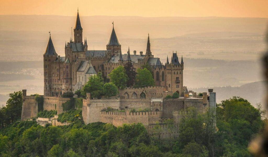Hohenzollern Castle in Germany perched atop a forested hill, featuring tall pointed towers and stone walls, with a hazy sunset landscape in the background.