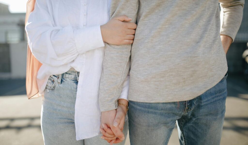 Close-up of a couple holding hands, with the woman’s arm linked around the man’s arm, both wearing casual jeans and long-sleeved tops.