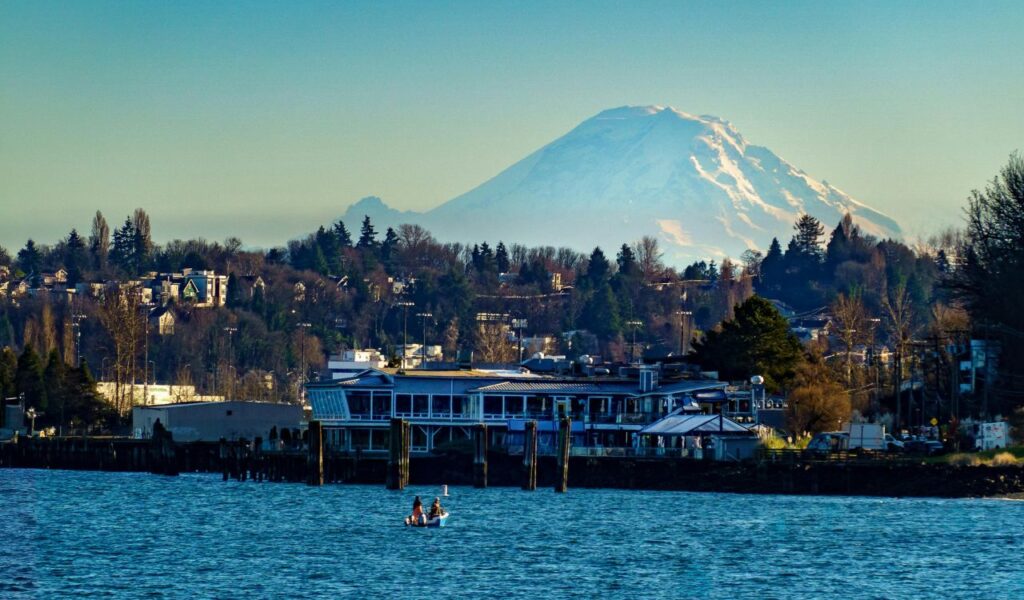 View of Hood River, Oregon, with waterfront buildings, a small boat on the river, forested hills, and snow-covered Mount Hood rising in the background.