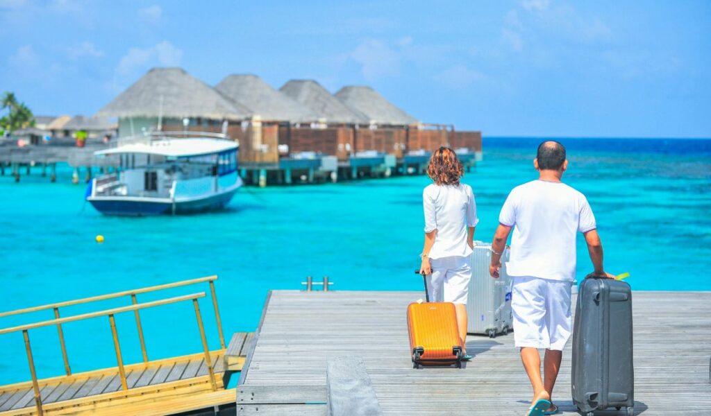 Two travelers walking on a wooden dock with suitcases toward overwater bungalows and a boat in a bright turquoise sea.