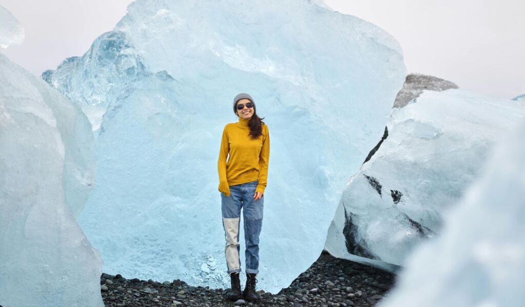 A smiling woman in a mustard sweater, jeans, boots, and sunglasses stands between massive blue ice formations on a rocky Icelandic shore.