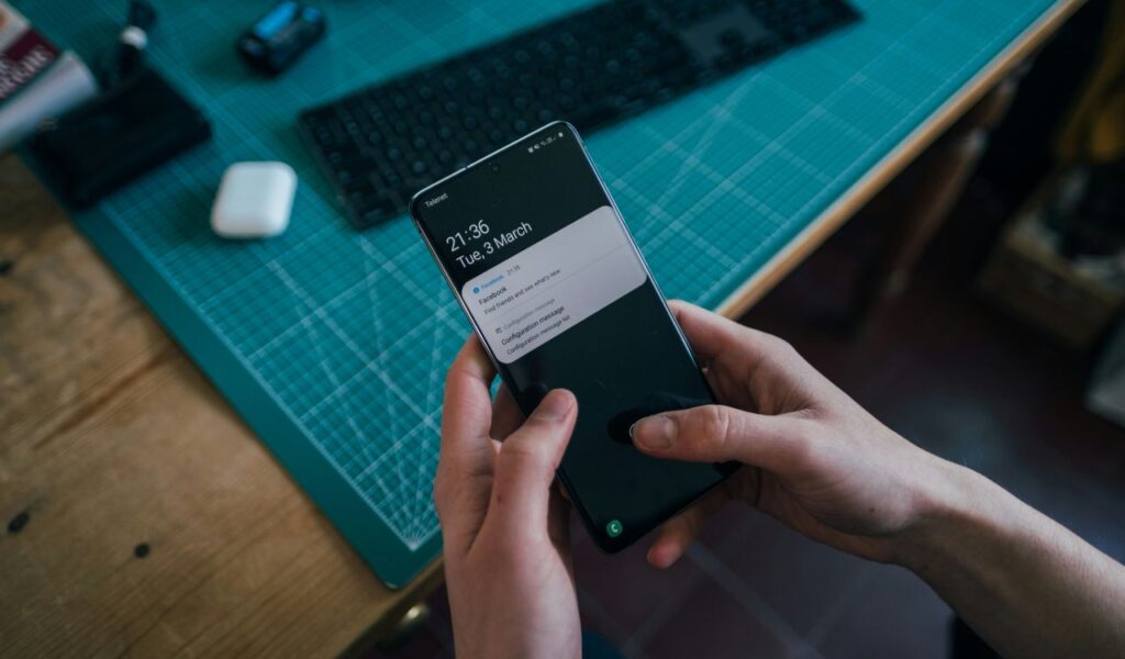 Person holding a smartphone displaying notifications while sitting at a desk with a keyboard and other items in the background.