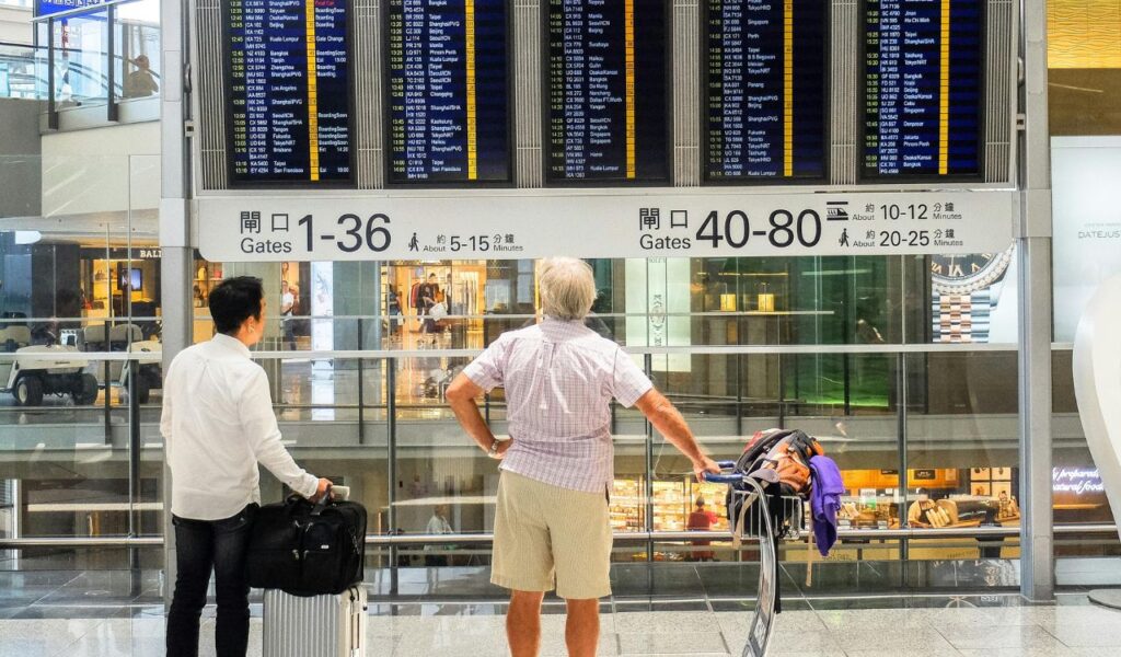 Two travelers check an airport departure board, luggage in hand, under signs showing gate directions and walking times.