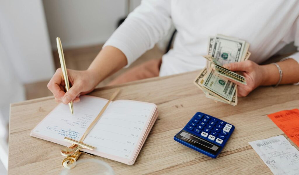 Person budgeting by writing in a planner while holding US dollar bills, with a blue calculator, receipts, and a gold pen on a wooden desk.