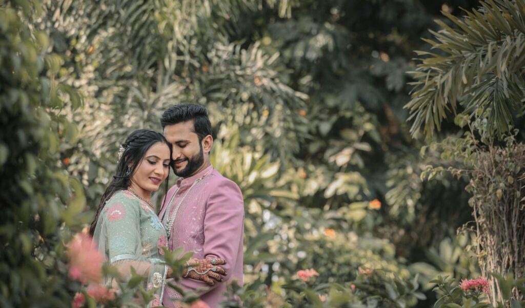  Couple in traditional attire embracing in a lush garden surrounded by greenery and flowers.