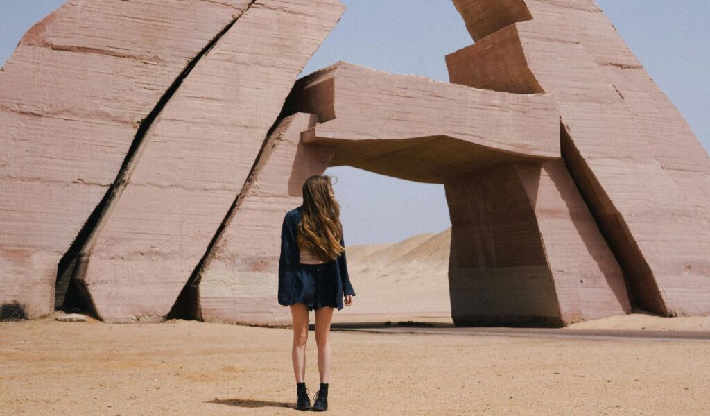 Woman standing in the desert facing large geometric stone structures forming an archway.