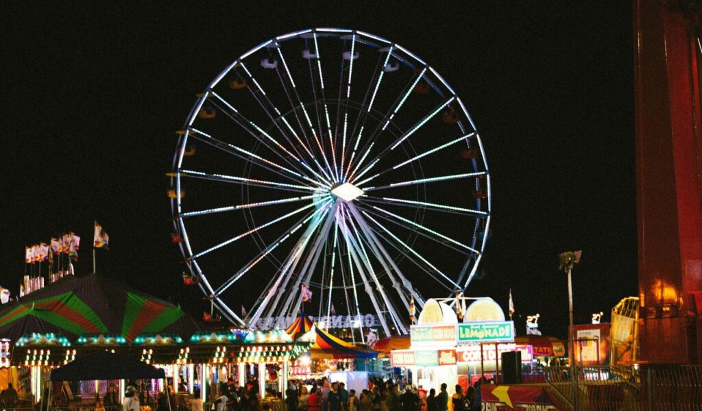 A brightly lit Ferris wheel towers over the Iowa State Fair at night, surrounded by colorful tents, food stalls, and bustling crowds.