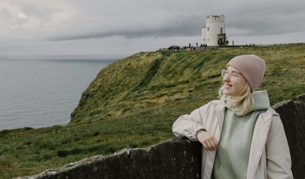 A smiling woman in a pink knit beanie, glasses, and a light jacket leans against a stone wall, with a grassy cliffside and a small tower overlooking the ocean in the background.