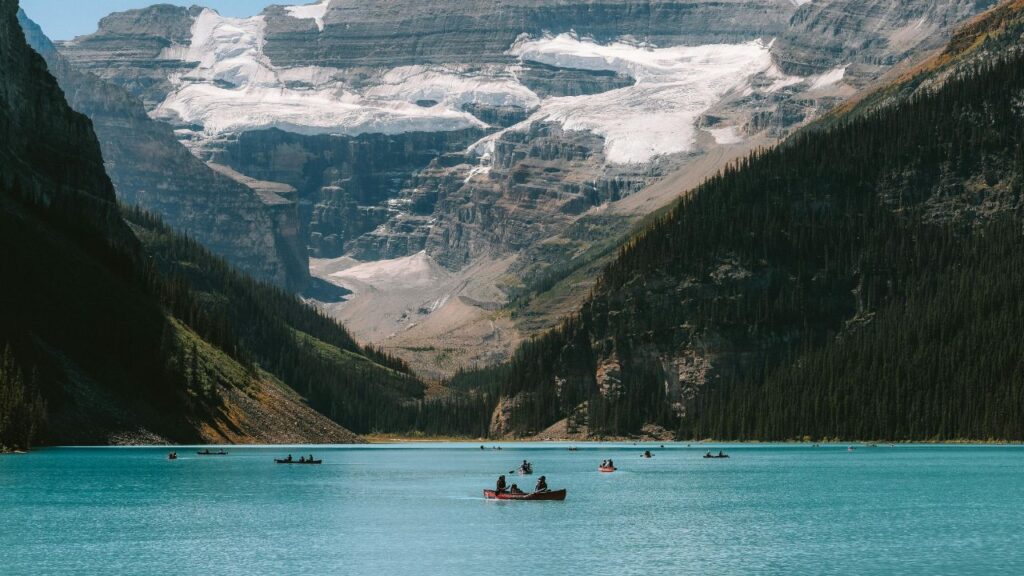  People paddling canoes on the turquoise waters of Lake Louise, surrounded by forested mountains and a backdrop of snow-covered glaciers in Banff National Park, Canada.