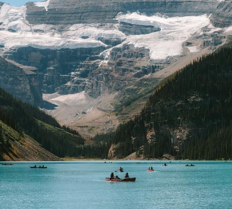 People paddling canoes on the turquoise waters of Lake Louise, surrounded by forested mountains and a backdrop of snow-covered glaciers in Banff National Park, Canada.