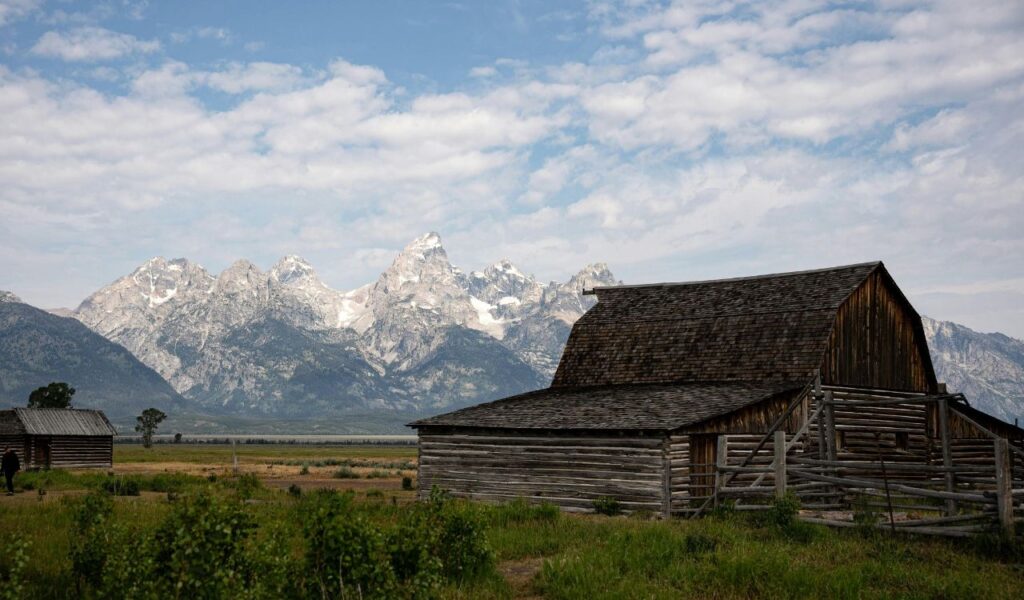 Historic wooden barn in Jackson Hole, Wyoming, set against a dramatic backdrop of the snow-capped Teton Mountain Range under a partly cloudy sky.