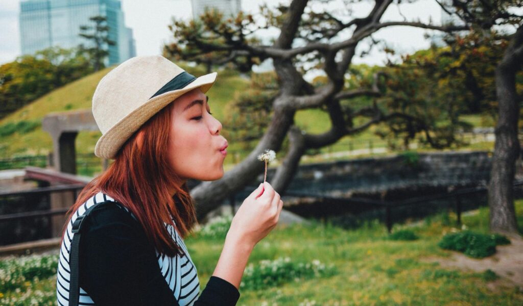 A woman in Japan enjoys a peaceful moment, blowing on a dandelion in a park with lush greenery and traditional landscaping in the background.