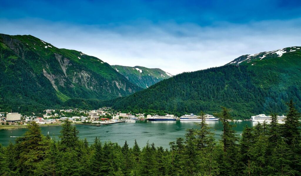 A scenic view of Juneau, Alaska, showing the city nestled between forested mountains and the harbor, with several cruise ships docked along the shoreline.