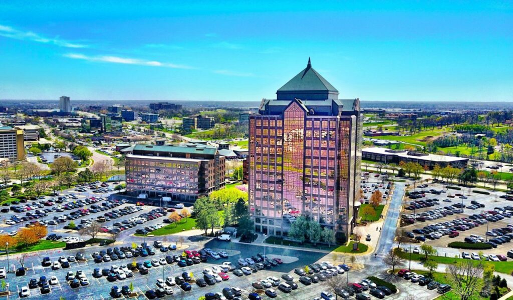 Aerial view of Kansas City, Missouri, featuring a tall office building with reflective glass windows, surrounded by parking lots filled with cars and a sprawling urban landscape under a clear blue sky