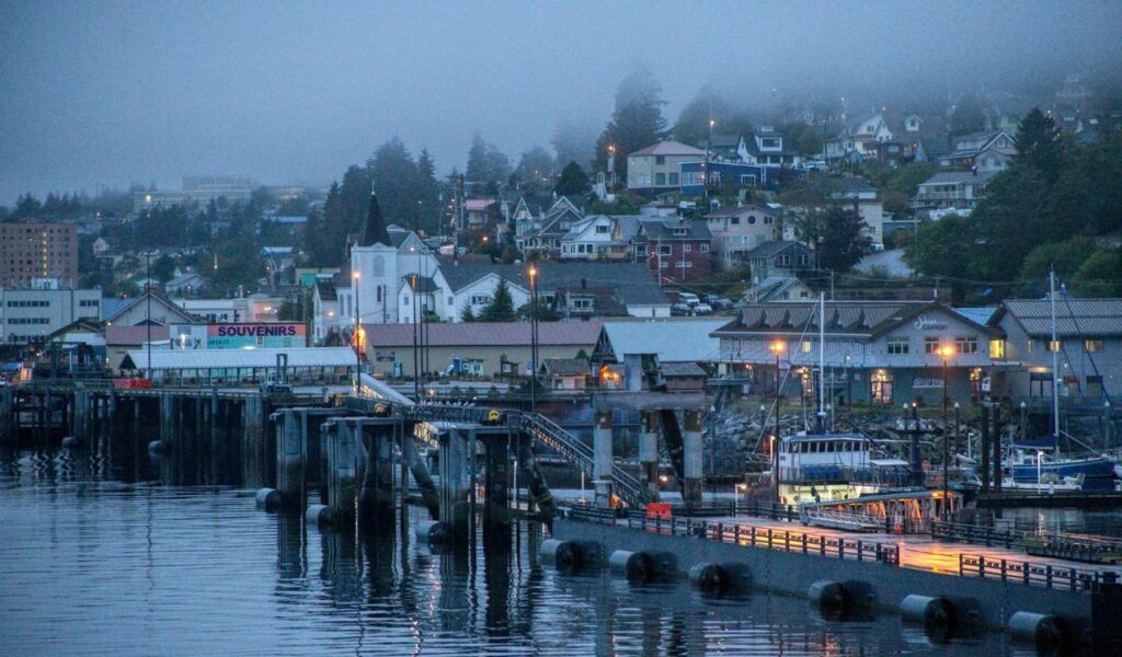 A foggy evening view of Ketchikan, Alaska, with waterfront docks, fishing boats, souvenir shops, and hillside homes illuminated by streetlights.
