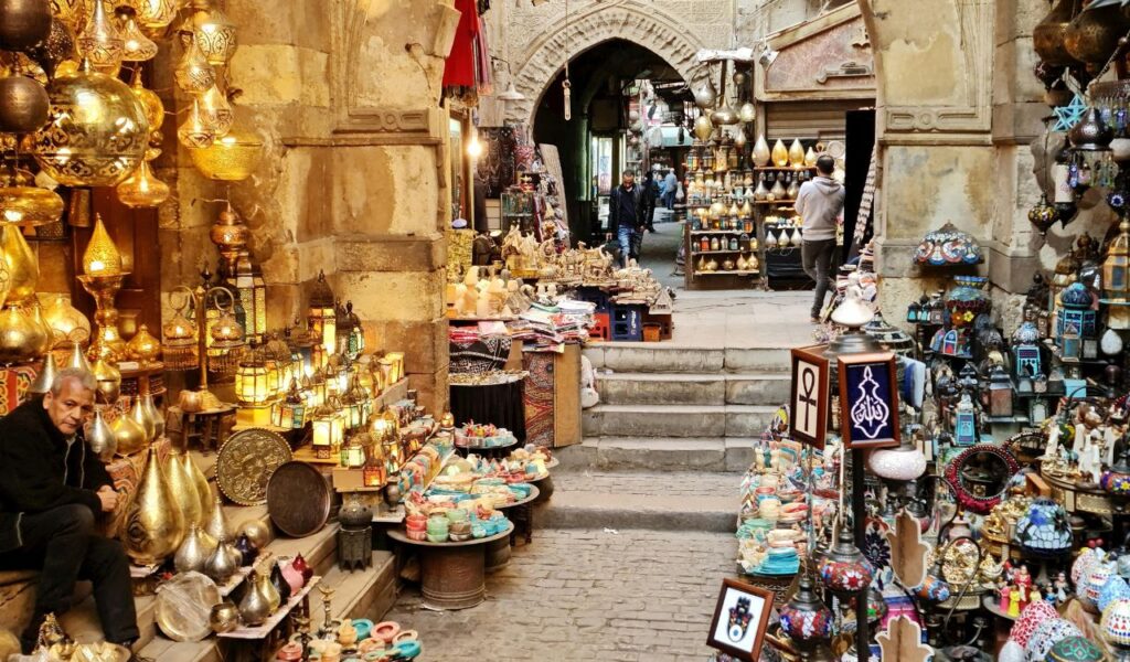 Narrow market street in Cairo filled with colorful lamps, pottery, and souvenirs, with shopkeepers and shoppers along the stone-paved path.