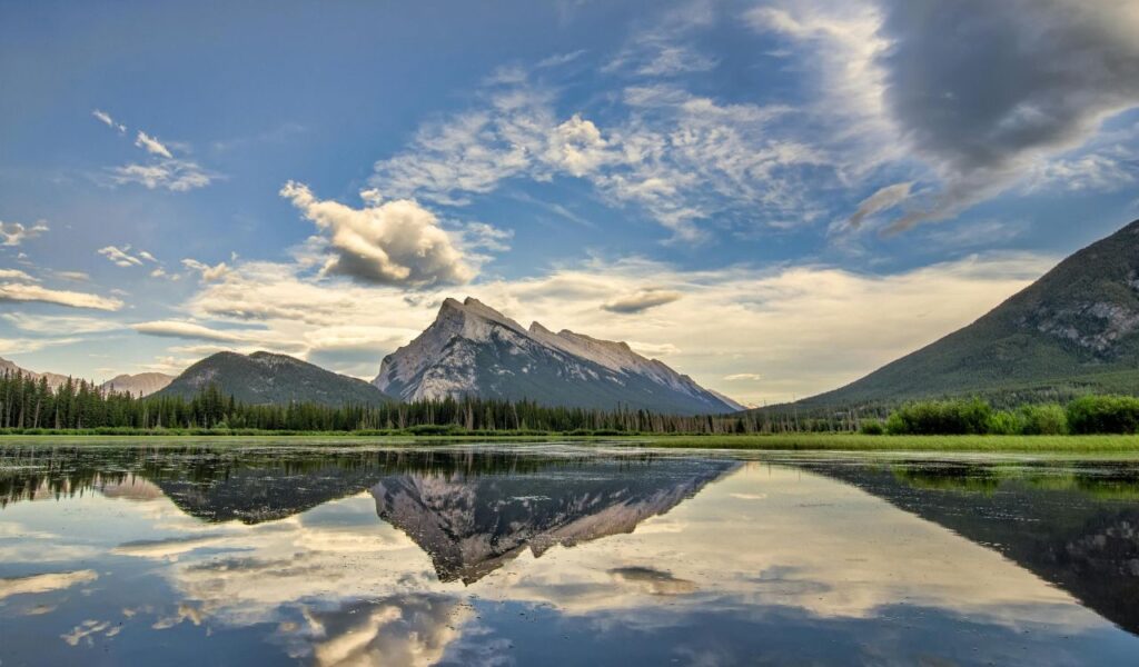 A dramatic mountain range reflects in the still waters of Lake Clark, Alaska, surrounded by green forests and under a sky of scattered clouds.