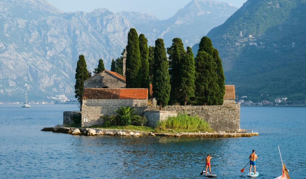 Paddleboarders glide past a small stone island with a red-roofed chapel and tall cypress trees, surrounded by Lake George and mountains.