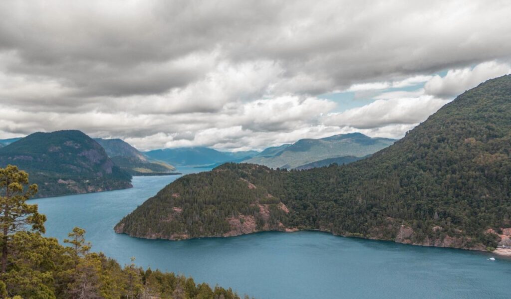 A winding blue lake curves through forested hills under a cloudy sky, with layers of mountains in the background and trees in the foreground.