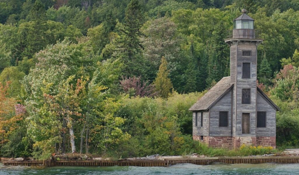 A weathered wooden lighthouse stands at the forested edge of Lake Superior, surrounded by dense trees and calm blue-green water.