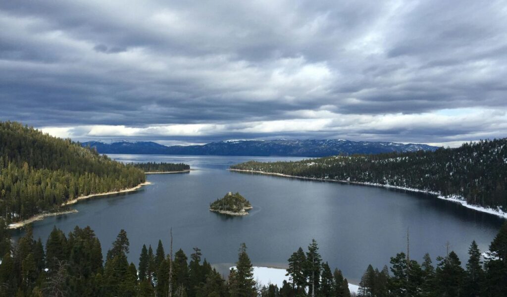 Scenic view of Lake Tahoe surrounded by pine-covered mountains, with a small island in the middle and cloudy skies above.