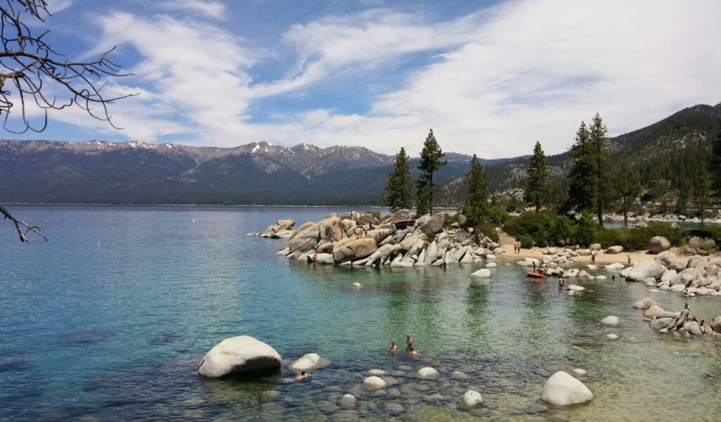 Clear turquoise water and smooth boulders line the shore of Lake Tahoe, with swimmers, pine trees, and snow-capped mountains in view.
