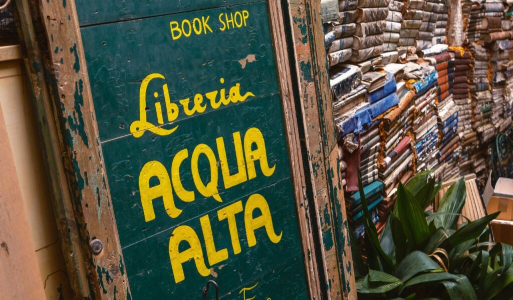 A weathered green sign reading "Libreria Acqua Alta" beside stacks of old books in an outdoor area with green plants.