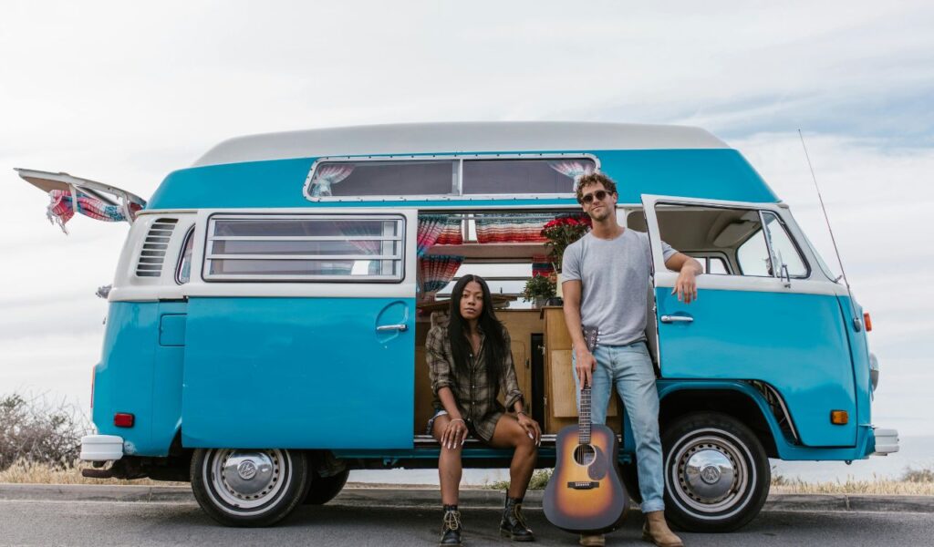 A man and woman on a road trip pose by a blue retro van with open doors, colorful curtains inside, and a guitar resting on the ground.