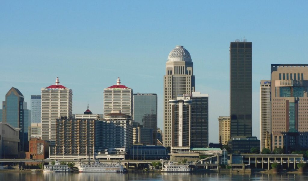 Skyline of Louisville, Kentucky, with tall modern buildings, a hotel, and riverboats docked along the waterfront under a clear blue sky