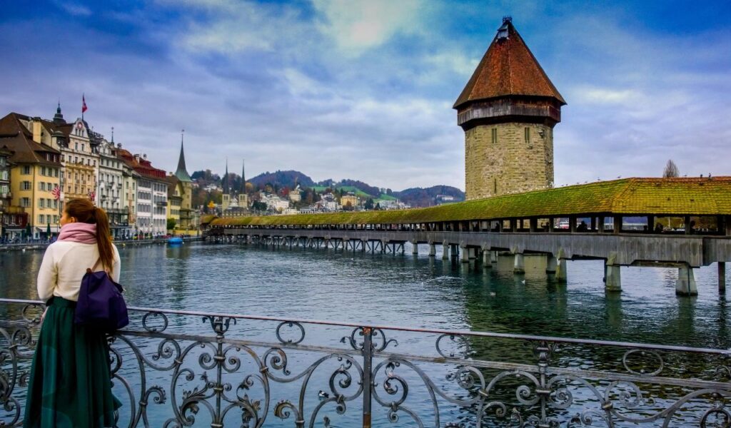 A woman stands by a railing overlooking Lucerne’s Chapel Bridge and Water Tower, with colorful buildings and hills in the background.