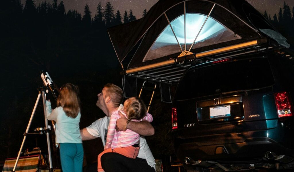 Father and daughters stargazing at a campsite with a rooftop tent on an SUV under a starry sky.