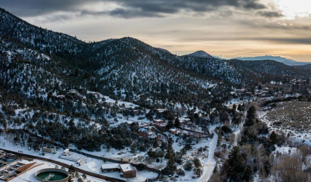 Aerial view of snow-covered hills and pine forests surrounding Santa Fe, New Mexico, with scattered houses, winding roads, and a dramatic winter sky at sunset