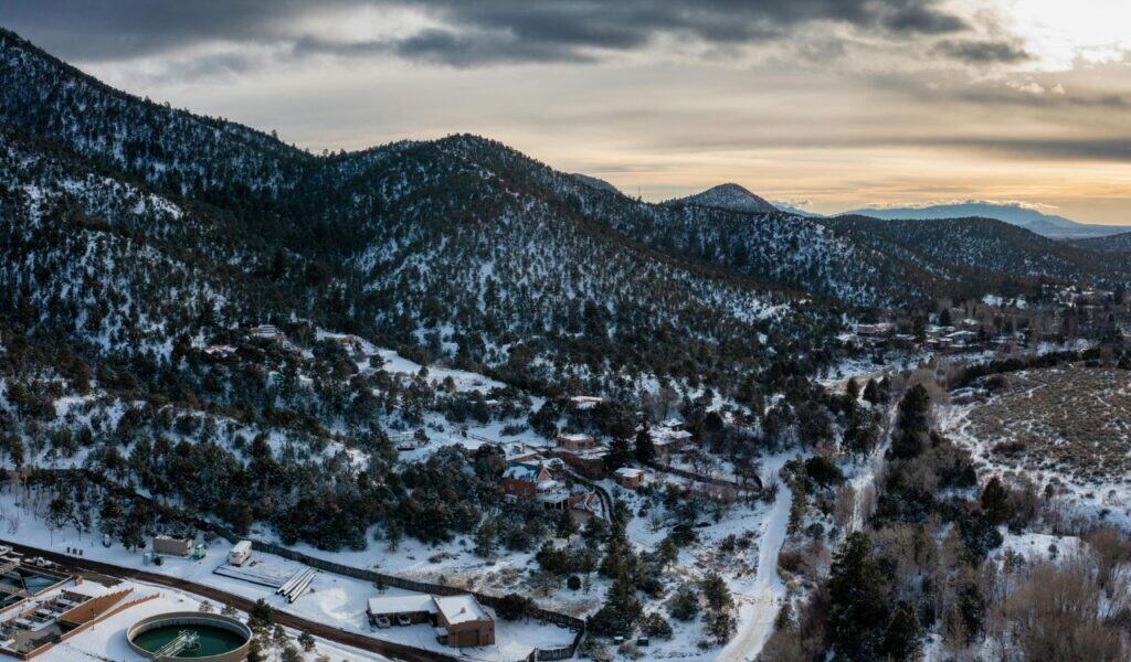 Aerial view of snow-covered hills and pine forests surrounding Santa Fe, New Mexico, with scattered houses, winding roads, and a dramatic winter sky at sunset.