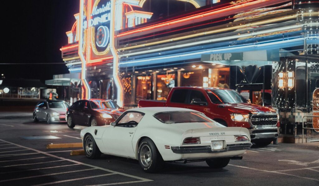 Night scene at Marietta Square in Marietta, Georgia, featuring a classic white Pontiac Trans Am parked outside a brightly lit diner with neon signs, alongside other vehicles in the parking lot.