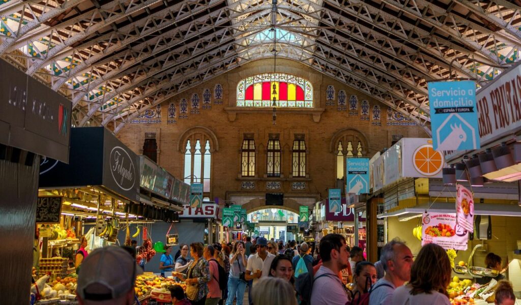 Crowds shopping inside Mercado Central, with high arched ceilings, stained glass windows, and stalls selling fresh produce.