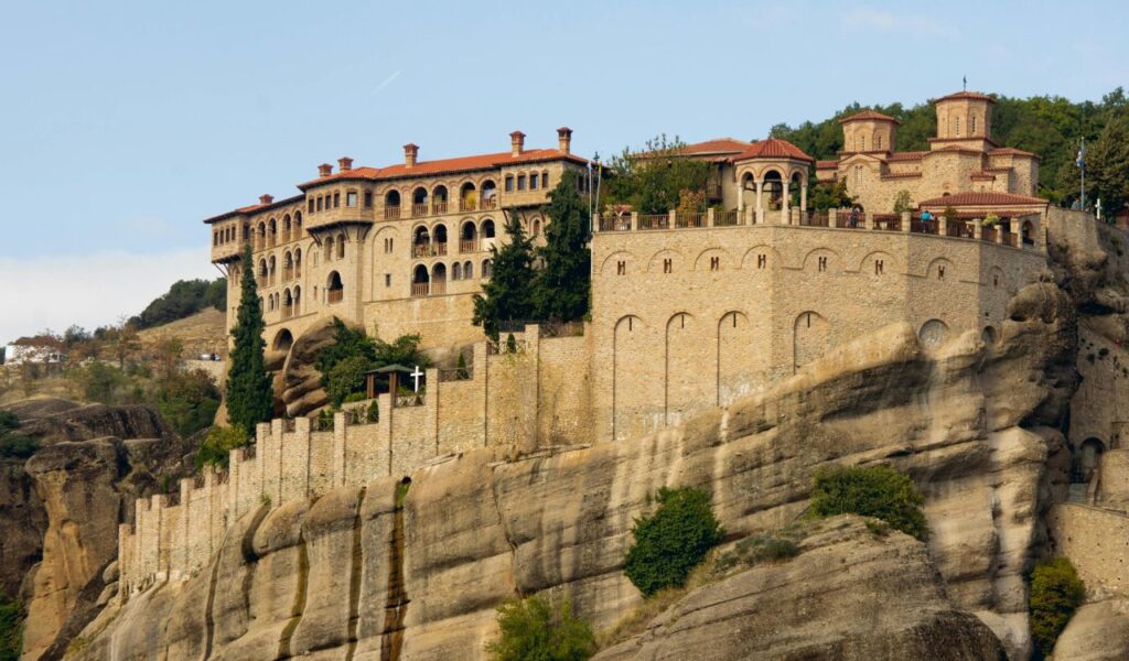 A historic Meteora monastery in Greece perched dramatically atop massive rock pillars, with stone walls blending into the rugged cliffs and lush greenery in the background.