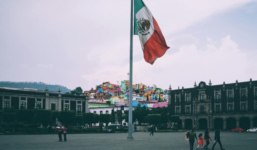 Large Mexican flag waving in Mexico City’s historic square with colonial buildings in the background and colorful hillside houses adding vibrant contrast.
