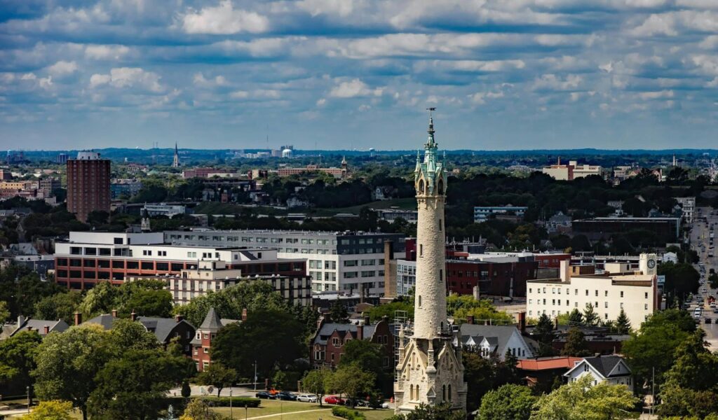 A tall, ornate stone water tower with a green copper roof stands in the foreground, surrounded by trees and historic houses, with the cityscape of Milwaukee and a partly cloudy sky in the background.
