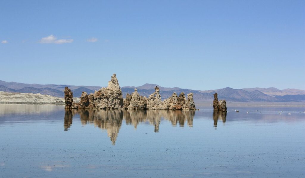 Tufa rock formations rise from the calm, reflective waters of Mono Lake in California, with distant mountains under a clear blue sky.