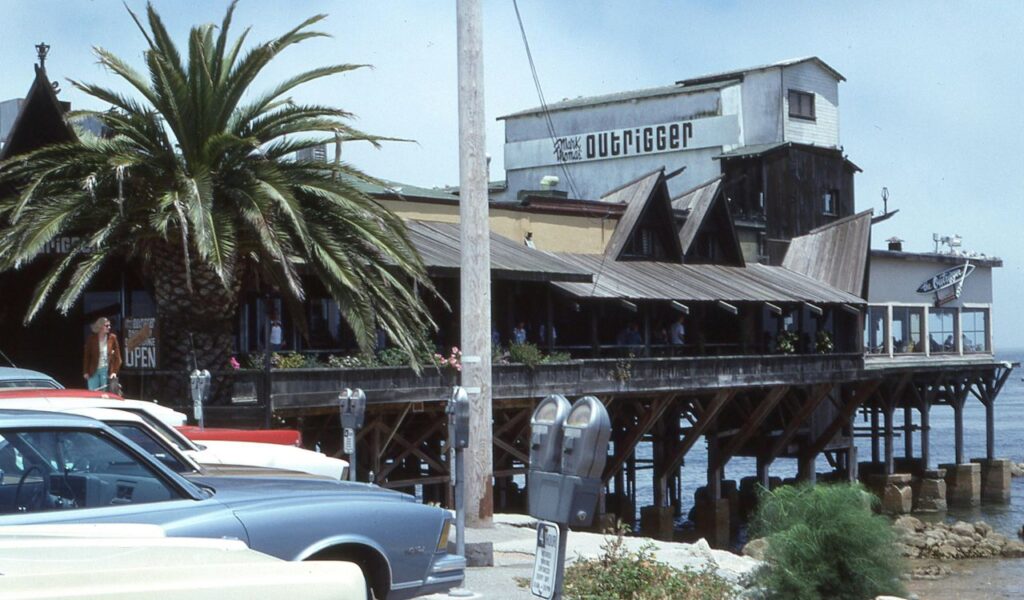 Vintage waterfront scene in Monterey, California, showing The Outrigger restaurant on wooden stilts above the ocean, with parked classic cars and a large palm tree in the foreground.