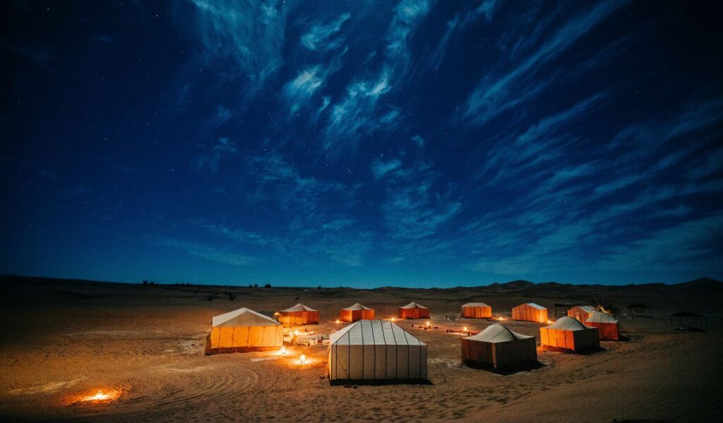 Tented desert camp glowing under a starry night sky with scattered campfires.
