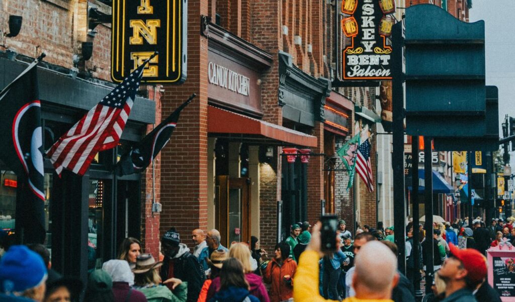 Crowded Nashville street with brick buildings, neon signs, and flags, featuring shops like Candy Kitchen and Whiskey Bent Saloon.