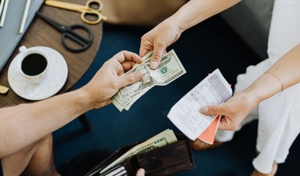 A close-up of hands exchanging US dollar bills and a receipt, with a wallet, scissors, and a cup of coffee on a nearby table.