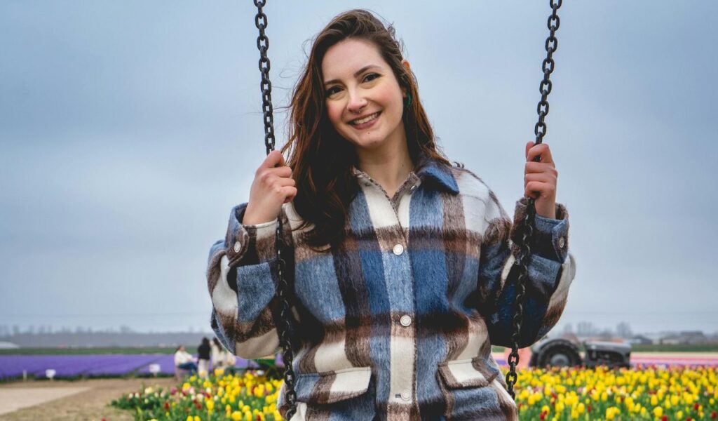 A smiling woman enjoys a swing in the Netherlands, surrounded by vibrant tulip fields and a soft overcast sky in the background.