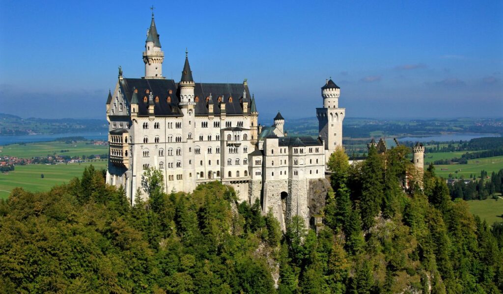 Neuschwanstein Castle perched on a forested hill in Bavaria, Germany, with its white towers and spires overlooking rolling green fields and distant lakes under a clear blue sky.