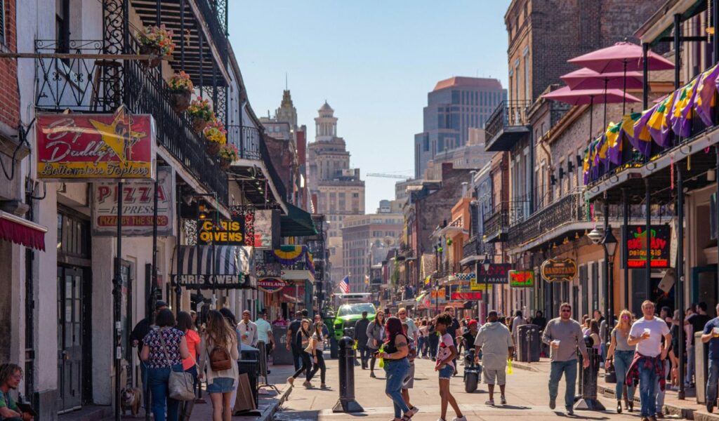  A lively street scene in New Orleans’ French Quarter with people walking, colorful signs, and historic buildings adorned with balconies and festive decorations.