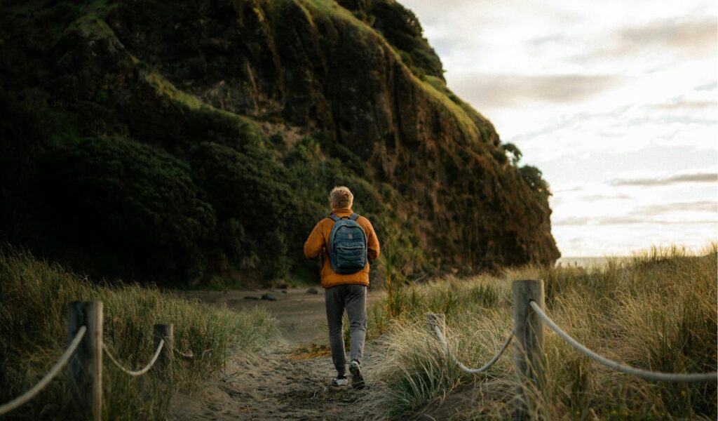 A lone traveler with a blue backpack walks along a sandy, grassy path toward a towering, green-clad cliff in New Zealand.